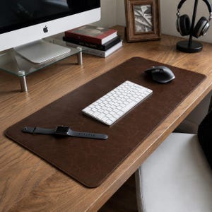 Puede incluir: Una alfombrilla de escritorio rectangular de color marrón oscuro con esquinas redondeadas y una superficie lisa y mate. La alfombrilla está colocada sobre un escritorio de madera y sirve de base para un teclado inalámbrico blanco, un ratón negro y un reloj inteligente negro.