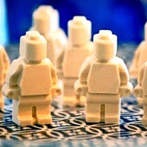May include: A group of seven off-white, unpainted plastic toy figures shaped like building block characters. Each figure features a cylindrical head with a stud on top, a rectangular torso, and articulated arms and legs, standing in a cluster.