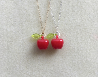 Puede incluir: Tres collares con colgantes de manzana roja y detalles de hojas verdes. Los collares están sobre un fondo beige claro. Las cadenas son doradas, plateadas y plateadas.