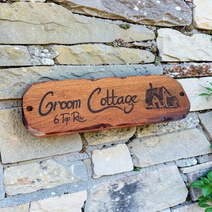 Pode incluir: Placa de madeira com as palavras "Groom Cottage" e "6 Top Row" gravadas, e uma ilustra&ccedil;&atilde;o de uma casa. A placa est&aacute; montada numa parede de pedra. A madeira &eacute; castanha.