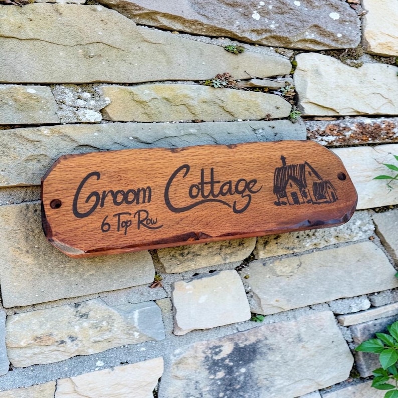 Pode incluir: Placa de madeira com as palavras "Groom Cottage" e "6 Top Row" gravadas, e uma ilustra&ccedil;&atilde;o de uma casa. A placa est&aacute; montada numa parede de pedra. A madeira &eacute; castanha.