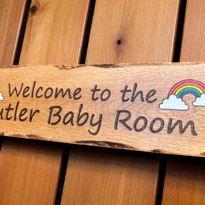 May include: A rectangular wooden sign with natural live edges featuring the engraved text Welcome to the Butler Baby Room. The sign includes two colorful rainbow and white cloud illustrations, one on each side of the text, and two small holes.
