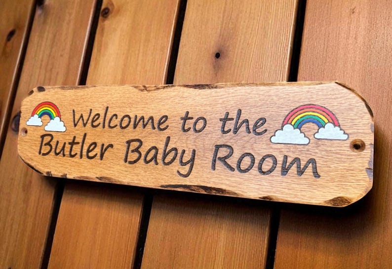 May include: A rectangular wooden sign with natural live edges featuring the engraved text Welcome to the Butler Baby Room. The sign includes two colorful rainbow and white cloud illustrations, one on each side of the text, and two small holes.