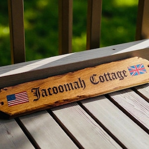 May include: A rectangular wooden sign with a natural live edge, featuring the engraved text "Jacoomah Cottage" in a dark font. The sign displays a small American flag on the left side and a British Union Jack flag on the right side, with pre-drilled holes.
