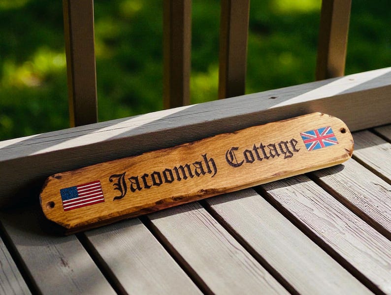 May include: A rectangular wooden sign with a natural live edge, featuring the engraved text "Jacoomah Cottage" in a dark font. The sign displays a small American flag on the left side and a British Union Jack flag on the right side, with pre-drilled holes.