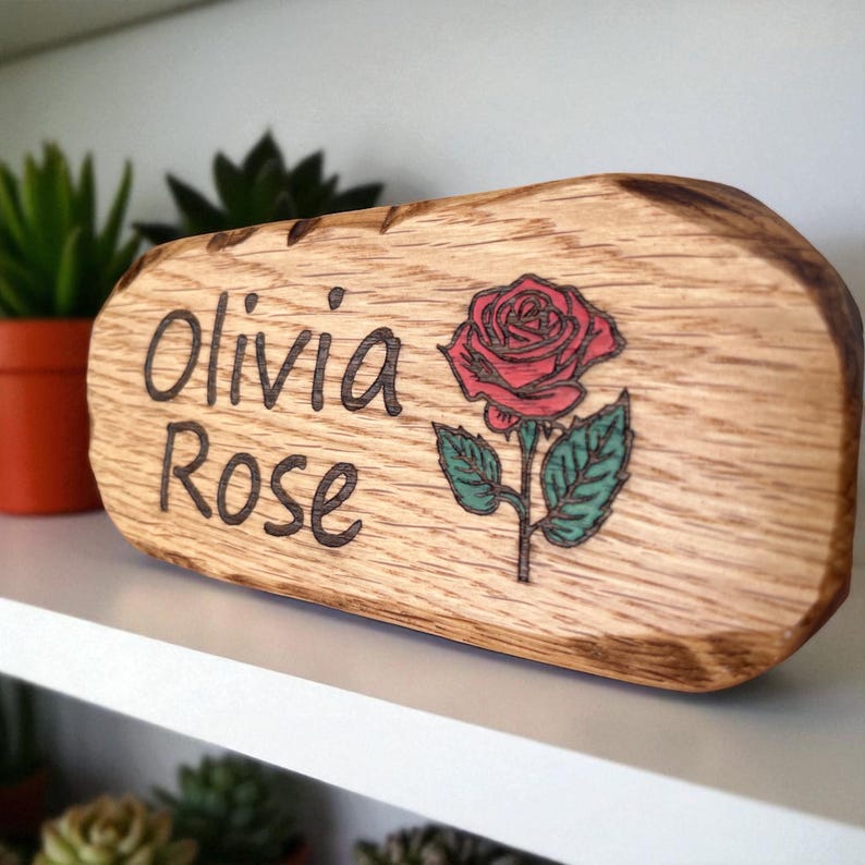 May include: A rectangular wooden plaque with rounded edges featuring the burned-in text Olivia Rose and a colored illustration of a red rose with green leaves. The wood grain is visible, and the edges have a dark, charred appearance.