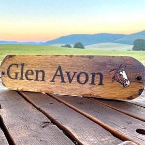 May include: A rectangular wooden sign with rounded edges featuring the text Glen Avon in black lettering and a graphic illustration of a brown horse head with a white blaze on its face. The sign has two small holes for mounting purposes.