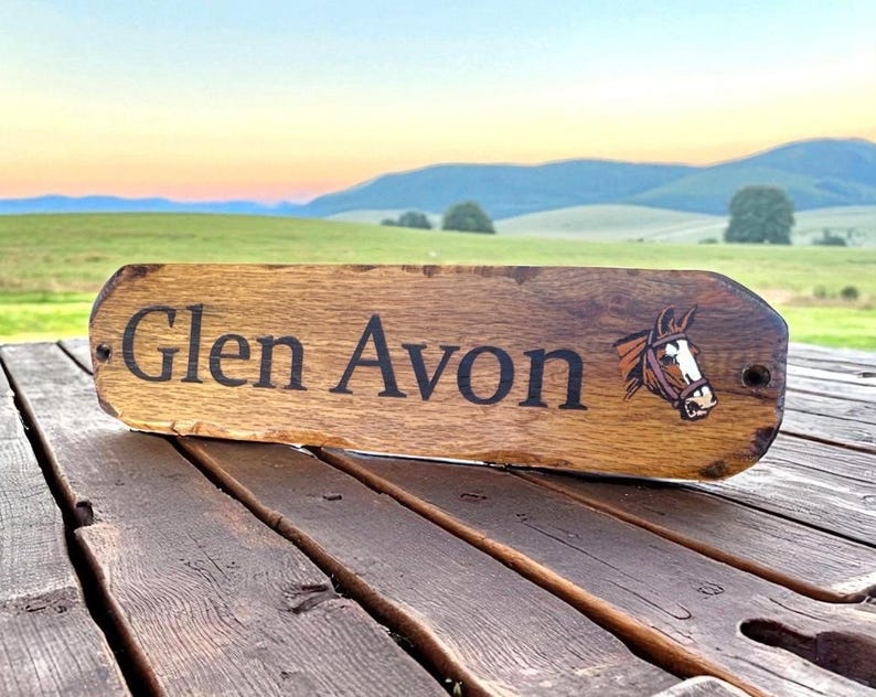 May include: A rectangular wooden sign with rounded edges featuring the text Glen Avon in black lettering and a graphic illustration of a brown horse head with a white blaze on its face. The sign has two small holes for mounting purposes.