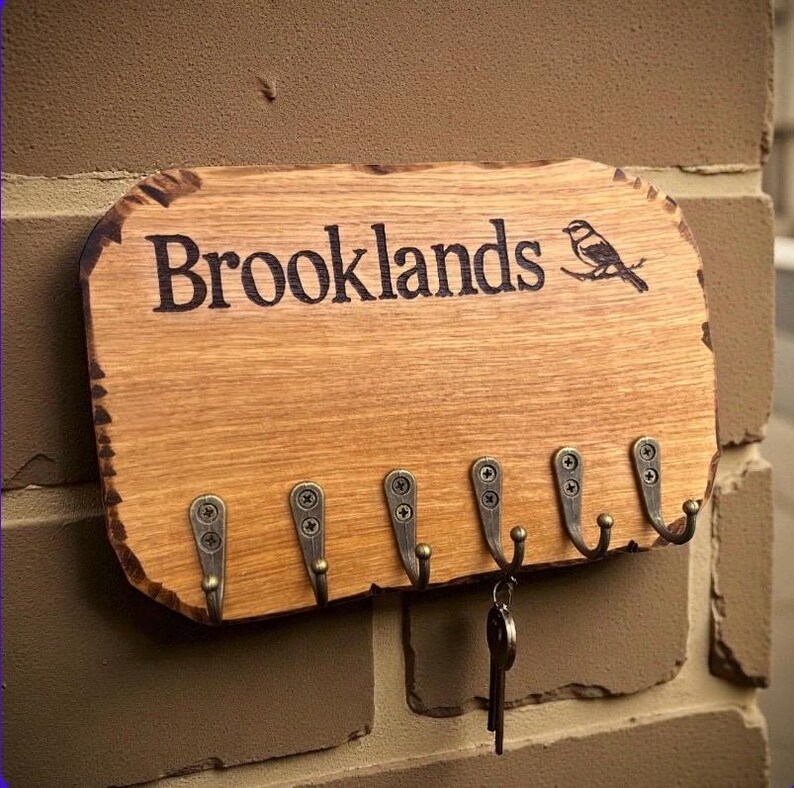 以下が含まれることがあります： 自然な縁を持つ長方形の木製キーホルダー。Brooklandsという刻印と鳥のイラストが施されています。ネジで固定された6つのアンティーク調の金属製フックが付いており、1つのフックに鍵の束が掛かっています。