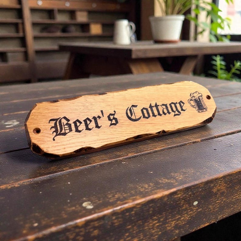 Pode incluir: Uma placa de madeira com o texto "Beer's Cottage" em uma fonte decorativa, acompanhada por uma ilustra&ccedil;&atilde;o de caneca de cerveja. A placa tem um gr&atilde;o de madeira natural e uma borda r&uacute;stica e irregular.