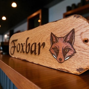 May include: A rectangular wooden sign with a natural live edge, featuring the word Foxbar engraved in a dark script font. To the right of the text is a detailed, colored illustration of a fox face with pointed ears and amber-colored eyes.