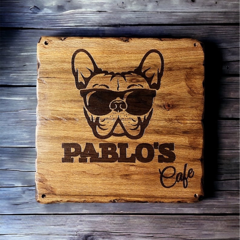 Pode incluir: Placa de madeira com uma imagem gravada de um buldogue franc&ecirc;s usando &oacute;culos de sol. A placa diz "PABLO'S Cafe". A placa quadrada tem bordas arredondadas e um acabamento natural de gr&atilde;o de madeira.