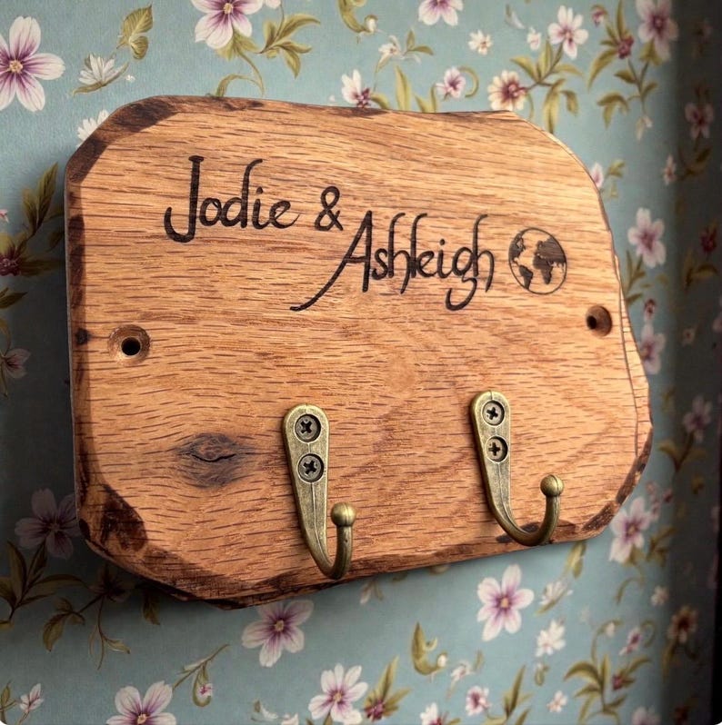 以下が含まれることがあります： Jodie & Ashleighという刻印と小さな地球儀のアイコンが施された、自然な縁を持つ長方形の木製プレート。アンティーク調の真鍮色をした金属製フック2つがネジで取り付けられており、取り付け用の穴が2つあけられています。