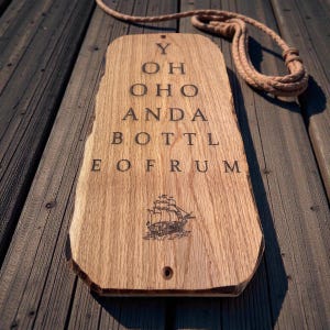 Pode incluir: Placa de madeira com o texto "YOHOHO AND A BOTTLE OF RUM" e uma ilustra&ccedil;&atilde;o de um navio. A placa &eacute; retangular com uma borda natural e um furo para pendurar. Uma corda est&aacute; presa no topo.