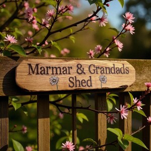 May include: A rectangular wooden sign with natural live edges featuring the engraved text "Marmar & Grandads Shed" in a dark font. Two small engraved daisy illustrations are positioned below the text on the light brown wood surface.