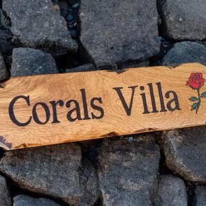 May include: A rectangular wooden sign with rounded edges and a natural live edge finish. The text Corals Villa is engraved in black lettering, accompanied by a small, detailed illustration of a red rose with green leaves on the right side.