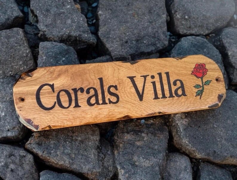 May include: A rectangular wooden sign with rounded edges and a natural live edge finish. The text Corals Villa is engraved in black lettering, accompanied by a small, detailed illustration of a red rose with green leaves on the right side.