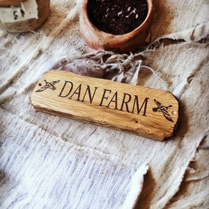 Pode incluir: Uma placa de madeira com o texto "DAN FARM" gravado em uma fonte serifada. A placa tem um gr&aacute;fico de pato em cada extremidade. A placa &eacute; retangular com bordas arredondadas. A placa &eacute; feita de madeira clara.