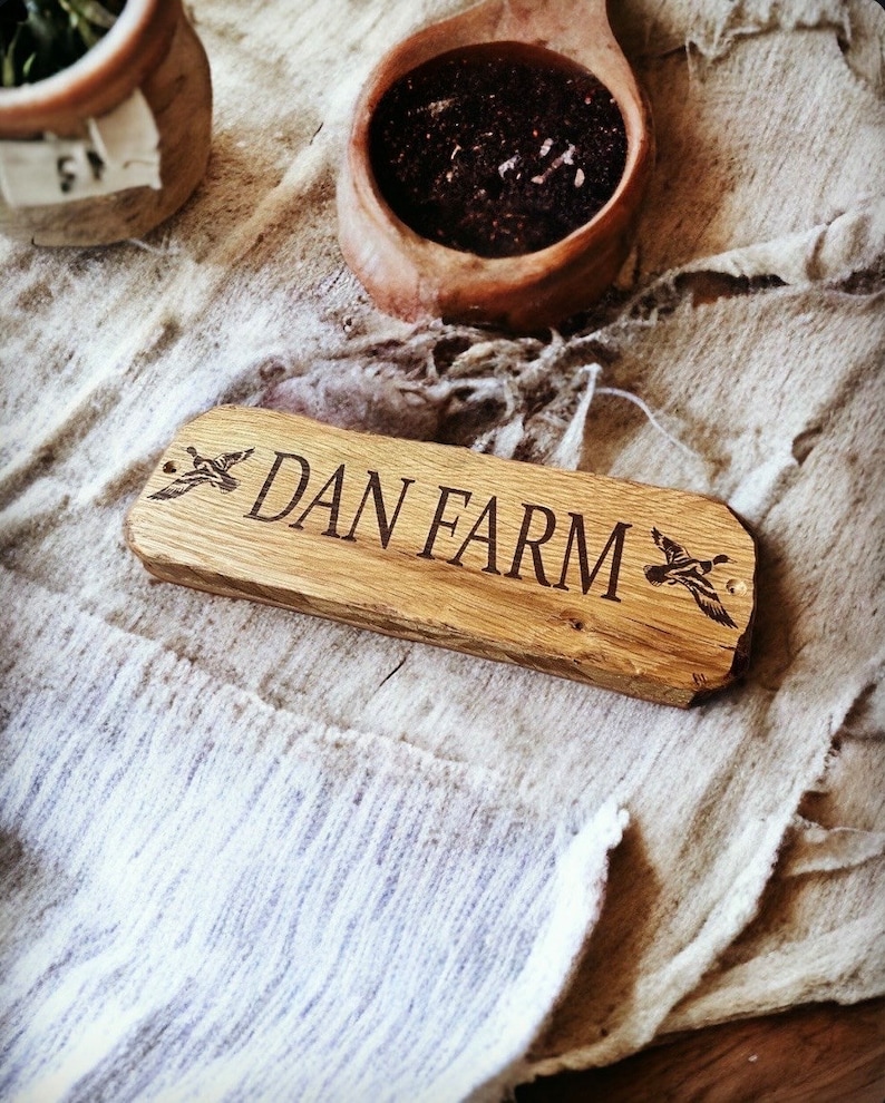 Pode incluir: Uma placa de madeira com o texto "DAN FARM" gravado em uma fonte serifada. A placa tem um gr&aacute;fico de pato em cada extremidade. A placa &eacute; retangular com bordas arredondadas. A placa &eacute; feita de madeira clara.