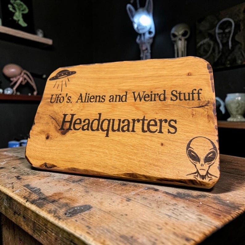 Pode incluir: Uma placa de madeira com o texto "Ufo's, Aliens and Weird Stuff Headquarters" &eacute; exibida. A placa apresenta uma imagem gravada de um OVNI e uma cabe&ccedil;a alien&iacute;gena. A madeira tem uma cor castanho-clara natural e uma borda &aacute;spera.
