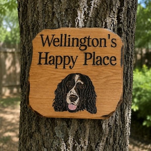 May include: A rectangular wooden sign with natural live edges featuring the engraved text "Wellington's Happy Place" in black lettering. Below the text is a detailed, colored illustration of a black and white spaniel dog with its tongue out.