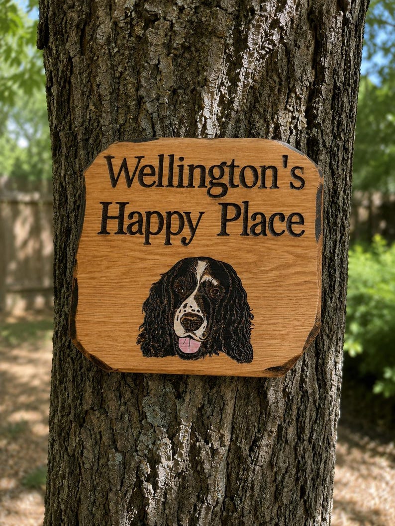 May include: A rectangular wooden sign with natural live edges featuring the engraved text "Wellington's Happy Place" in black lettering. Below the text is a detailed, colored illustration of a black and white spaniel dog with its tongue out.