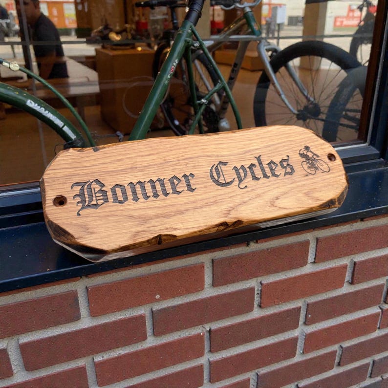 Pode incluir: Uma placa de madeira com as palavras "Bonner Cycles" em uma fonte ornamentada, juntamente com um pequeno gr&aacute;fico de bicicleta. A placa &eacute; de cor marrom claro e tem uma borda natural inacabada. &Eacute; exibida em frente a uma janela, com uma bicicleta vis&iacute;vel ao fundo.