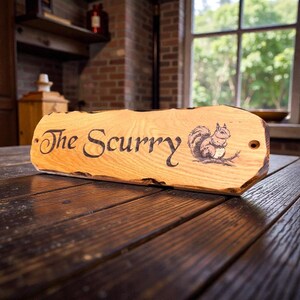 May include: A rectangular wooden sign with a natural live edge, featuring the text "The Scurry" in a dark script font and a detailed illustration of a squirrel on the right side. The sign has two pre-drilled holes on the left and right ends.