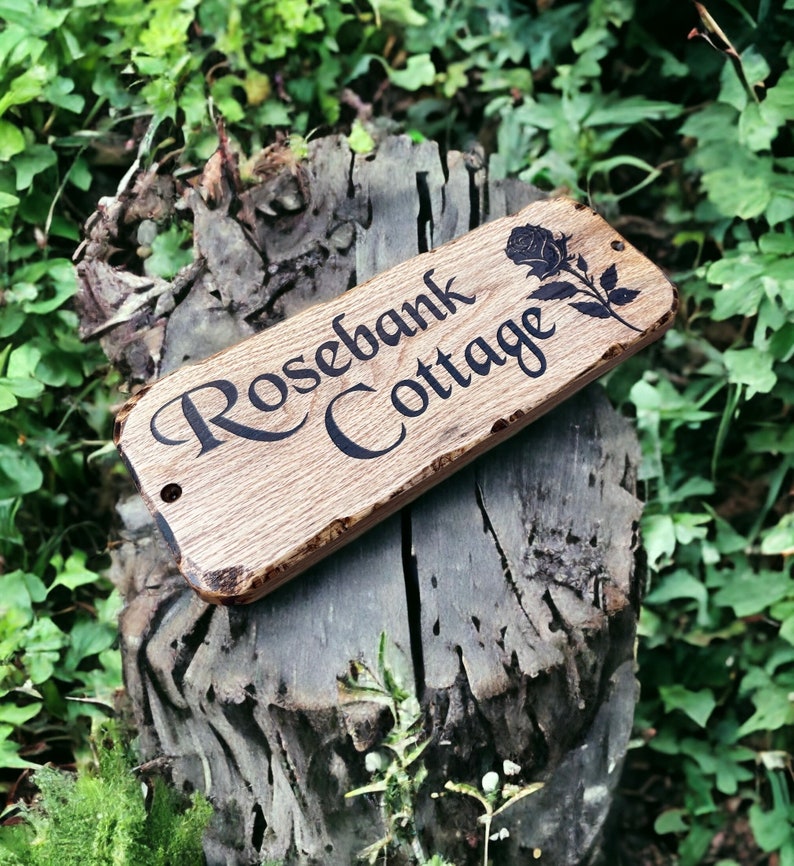 Pode incluir: Placa de madeira com as palavras "Rosebank Cottage" em uma fonte decorativa, acompanhada por uma ilustra&ccedil;&atilde;o de rosa. A placa &eacute; retangular com gr&atilde;o de madeira natural e uma borda r&uacute;stica. &Eacute; montada em um toco de &aacute;rvore desgastado.