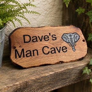 May include: A rectangular wooden sign with a natural live edge, featuring the engraved black text "Dave's Man Cave" and a detailed illustration of a grey elephant head on the right side. The sign has two small circular holes for mounting.