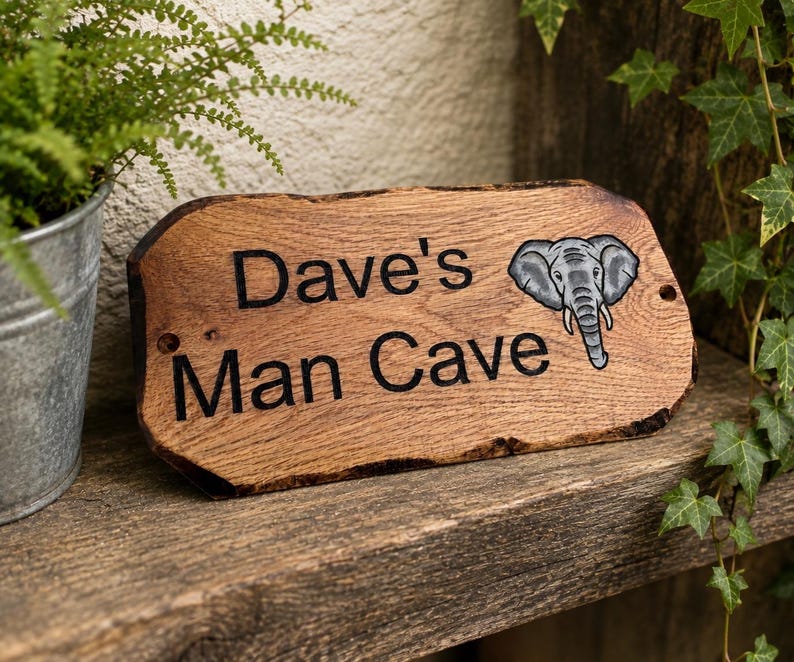 May include: A rectangular wooden sign with a natural live edge, featuring the engraved black text "Dave's Man Cave" and a detailed illustration of a grey elephant head on the right side. The sign has two small circular holes for mounting.