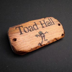May include: A rectangular wooden sign with natural live edges and two pre-drilled holes. The surface features the laser-engraved text "Toad Hall" in a serif font, accompanied by a small, colorful illustration of a standing toad character.