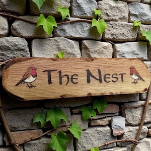 May include: A rectangular wooden sign with natural live edges featuring the text "The Nest" in dark, carved lettering. The sign is decorated with a small, hand-painted robin bird on both the left and right sides of the central text.