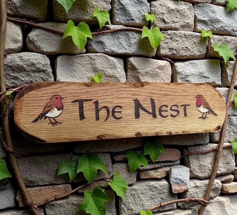 May include: A rectangular wooden sign with natural live edges featuring the text "The Nest" in dark, carved lettering. The sign is decorated with a small, hand-painted robin bird on both the left and right sides of the central text.