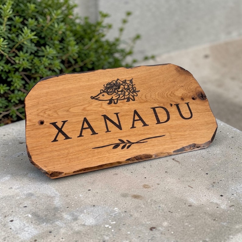 Pode incluir: Uma placa de madeira com a palavra "XANADU" em letras pretas. Acima do texto, h&aacute; um desenho de um ouri&ccedil;o e abaixo, um pequeno desenho de folhas. A placa tem um gr&atilde;o de madeira natural e uma borda r&uacute;stica inacabada.