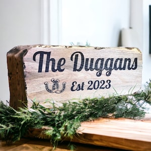 Pode incluir: Uma placa de madeira r&uacute;stica com o texto "The Duggans Est 2023" em preto. A placa apresenta um design de ferradura e &eacute; decorada com vegeta&ccedil;&atilde;o. A placa &eacute; colocada sobre uma superf&iacute;cie de madeira.