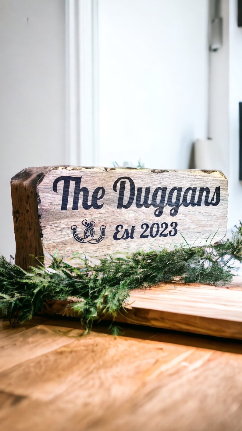 Pode incluir: Uma placa de madeira r&uacute;stica com o texto "The Duggans Est 2023" em preto. A placa apresenta um design de ferradura e &eacute; decorada com vegeta&ccedil;&atilde;o. A placa &eacute; colocada sobre uma superf&iacute;cie de madeira.