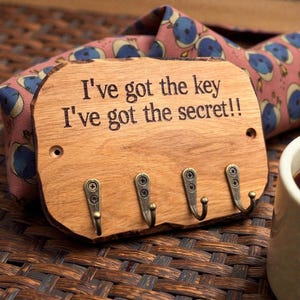 以下が含まれることがあります： 角が丸い長方形の木製キーホルダー。表面には「I've got the key I've got the secret!!」という文字が刻印されており、鍵や小物を掛けるためのアンティーク調の真鍮色金属製フックが4つ、ネジで取り付けられています。