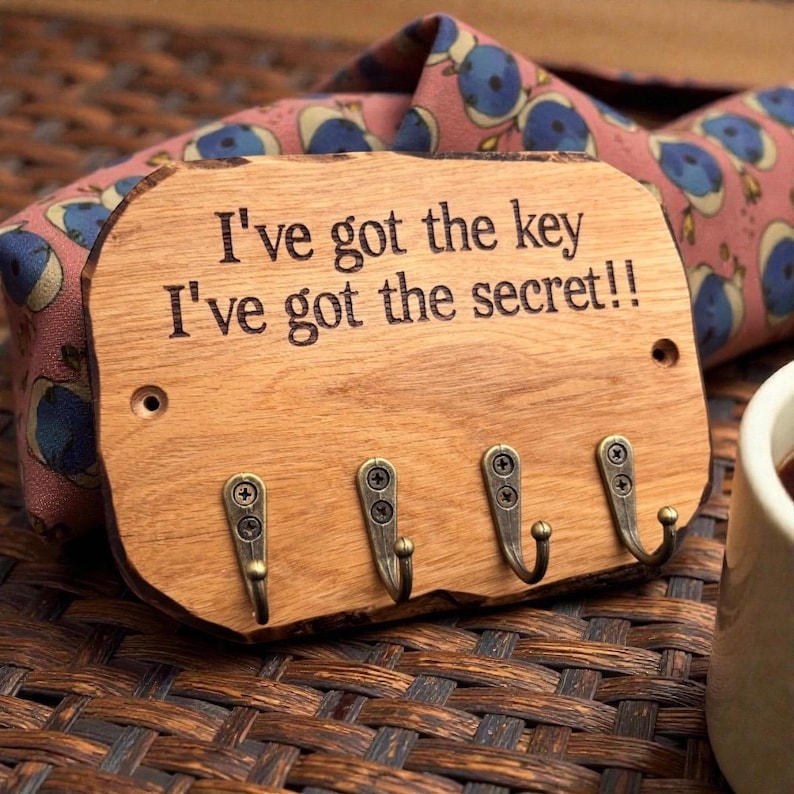 以下が含まれることがあります： 角が丸い長方形の木製キーホルダー。表面には「I've got the key I've got the secret!!」という文字が刻印されており、鍵や小物を掛けるためのアンティーク調の真鍮色金属製フックが4つ、ネジで取り付けられています。