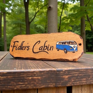 May include: A rectangular wooden sign with a natural live edge, featuring the text "Fidler's Cabin" in black script lettering and a colorful illustration of a blue and white vintage camper van on the right side, with two small mounting holes.
