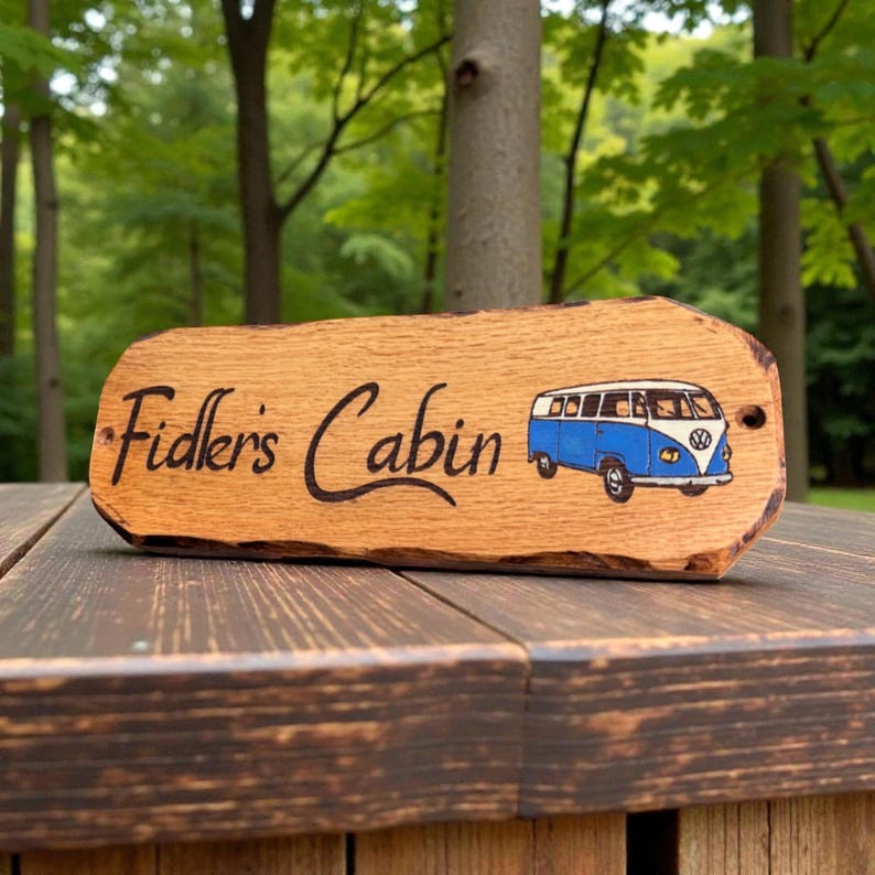 May include: A rectangular wooden sign with a natural live edge, featuring the text "Fidler's Cabin" in black script lettering and a colorful illustration of a blue and white vintage camper van on the right side, with two small mounting holes.