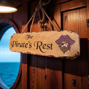 May include: A rectangular wooden sign with rough, dark edges hanging by a thick rope. The sign features the text "The Pirate's Rest" in a dark, serif font and a colorful illustration of a bearded pirate wearing a red bandana and an eye patch.