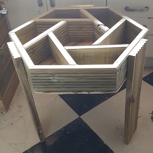 May include: A wooden hexagonal planter box with angled legs. The planter is constructed from unfinished wood and features internal dividers, creating multiple planting sections. The design is suitable for a garden or patio setting.