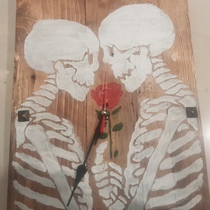 Könnte beinhalten: Wanduhr aus Holz mit einem gemalten Design von zwei weißen Skeletten, die sich gegenüberstehen, mit einer roten Rose dazwischen. Die Uhr hat schwarze Zeiger und eine goldfarbene Mitte. Die Uhr ist mit vier quadratischen Metallstiften befestigt.