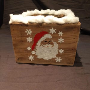 Könnte beinhalten: Eine Holzkiste mit einem weißen Baumwollrand, bemalt mit einem weißen Weihnachtsmann-Gesicht und einem roten Hut, sowie weißen Schneeflocken.