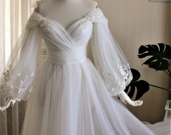 May include: A white off-the-shoulder wedding dress with long, sheer sleeves. The bodice features delicate floral embellishments and a wrapped waist. The skirt flows gracefully, creating an elegant and romantic silhouette. The dress is displayed on a mannequin.