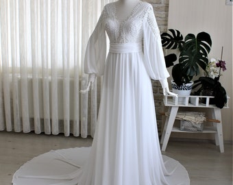 May include: A white wedding dress with a V-neckline and long, flowing sleeves. The bodice features intricate lace detailing, and the dress has a flowing skirt with a train. The dress is displayed on a mannequin, showcasing its elegant design.