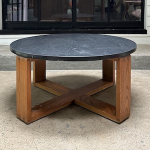 May include: A round coffee table with a dark grey stone top and a wooden base. The base features a cross-shaped design and is made of light brown wood. The table is approximately 76 cm in diameter.