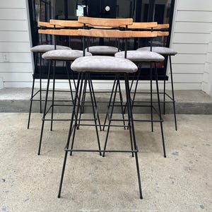 May include: Six bar stools with a mid-century modern aesthetic. They have light grey upholstered seats, light-coloured wooden backrests, and black metal frames. The stools are arranged in a row, highlighting their design and construction, suitable for a kitchen or bar.