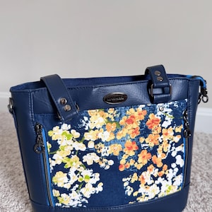 Puede incluir: Un bolso tote azul marino con un diseño floral. El bolso presenta un bolsillo frontal con un colorido estampado floral en amarillo, naranja y blanco. Tiene dos bolsillos laterales con cremalleras y dos asas. El bolso está hecho de un material duradero.